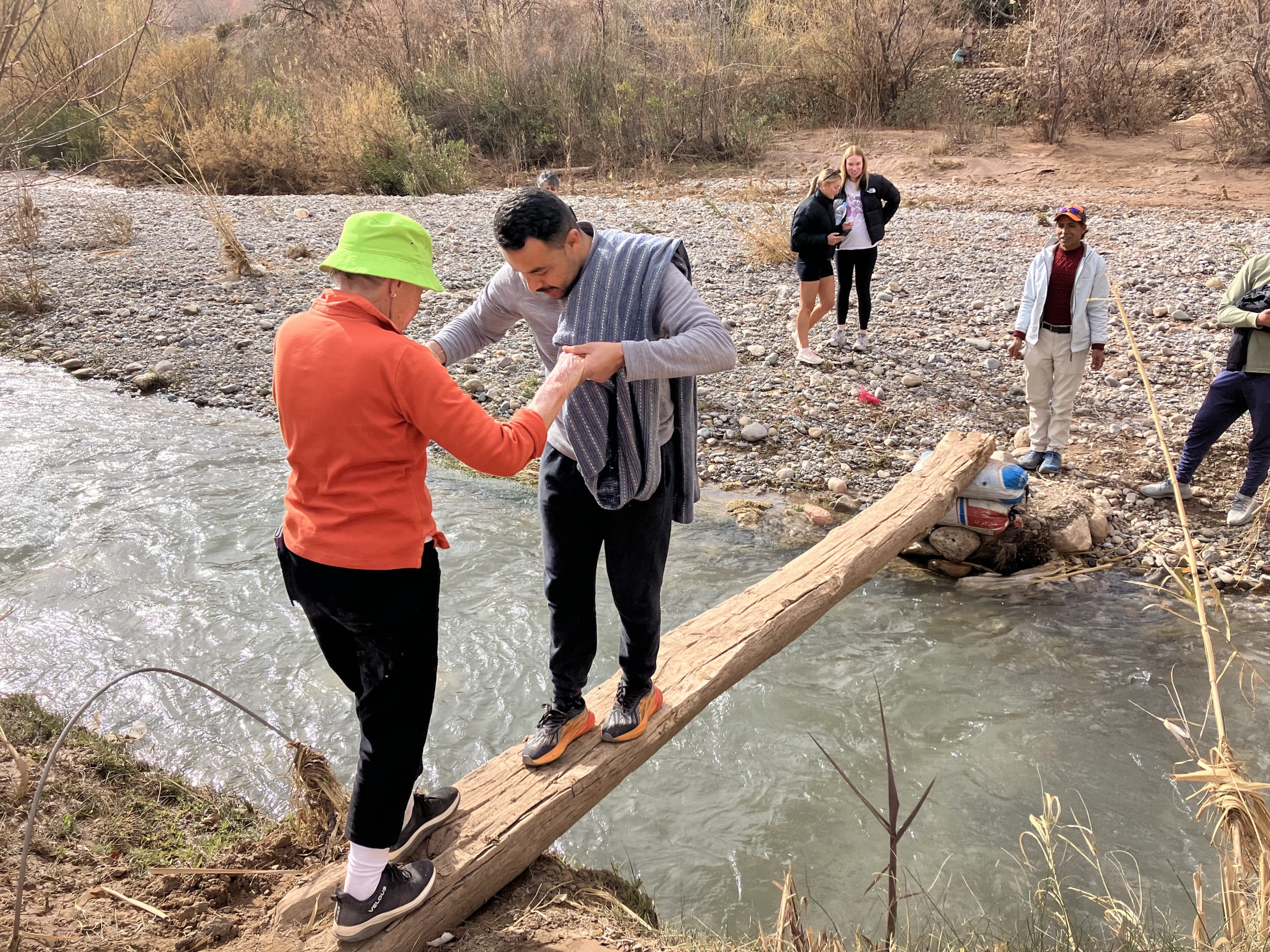 Hasy holding a guest's hand as she crosses a narrow wooden bridge over a stream in the Atlas Mountains